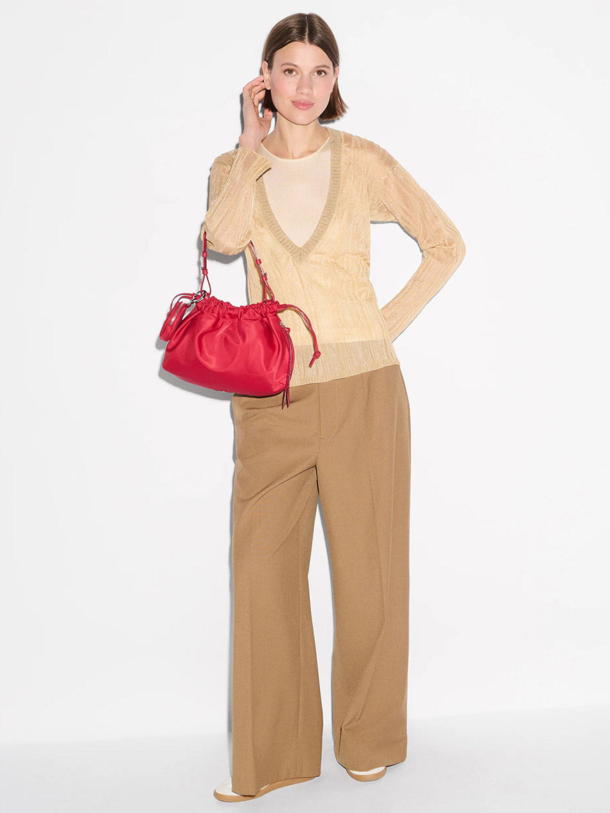 A woman stands against a white background wearing a beige sweater, wide-leg brown pants, and holding the MZ Wallace Small Waverly Shoulder in Apple Bedford, a bright red Italian leather bag with a magnetic closure.
