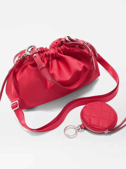 The MZ Wallace Small Waverly Shoulder in Apple Bedford is a red crossbody bag with Italian leather, drawstring and magnetic closure, and a detachable quilted coin pouch on the strap, displayed against a plain white background.