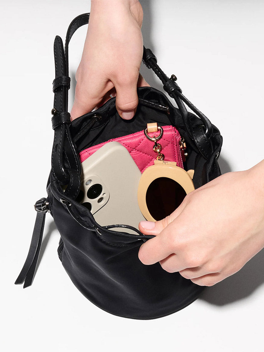 A person places a compact mirror into an MZ Wallace Waverly Bucket Bag in Black Bedford, which already holds a smartphone and a pink wallet.