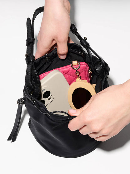 A person places a compact mirror into an MZ Wallace Waverly Bucket Bag in Black Bedford, which already holds a smartphone and a pink wallet.