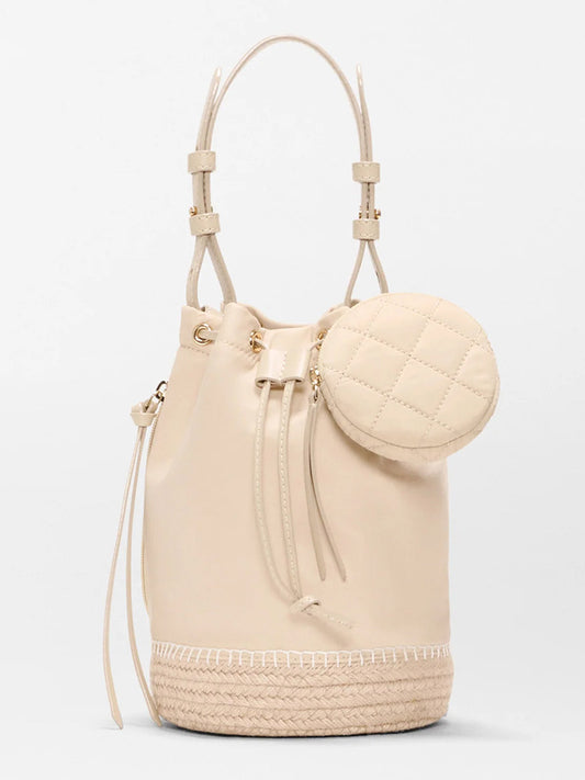 The MZ Wallace Waverly Espadrille Bucket Bag in Buff Bedford features a beige design with a drawstring closure, Italian leather trim, a round quilted coin pouch, and a braided base, shown against a plain white background.