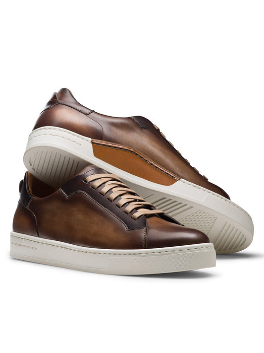 The Magnanni Amadeo in Taupe & Brown features brown leather cupsole sneakers with white soles; one shoe stands upright, while the other—crafted in Spain—is propped against it with its sole facing outward.