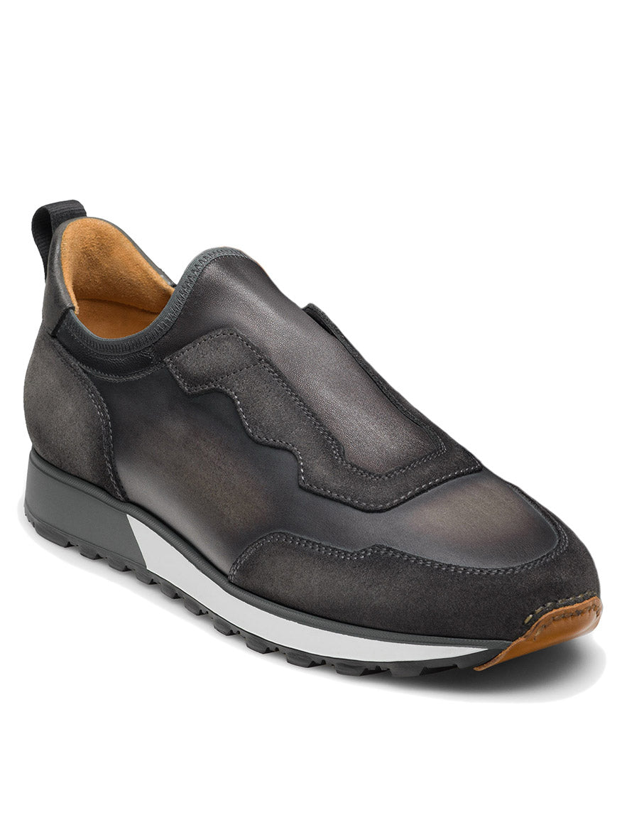 The Magnanni Arco Slip in Grey is a retro-inspired slip-on sport sneaker featuring a dark grey design with a white midsole, black outsole, and tan accents on the toe and heel. Its stretch-napa gore system ensures easy wearability and comfort.