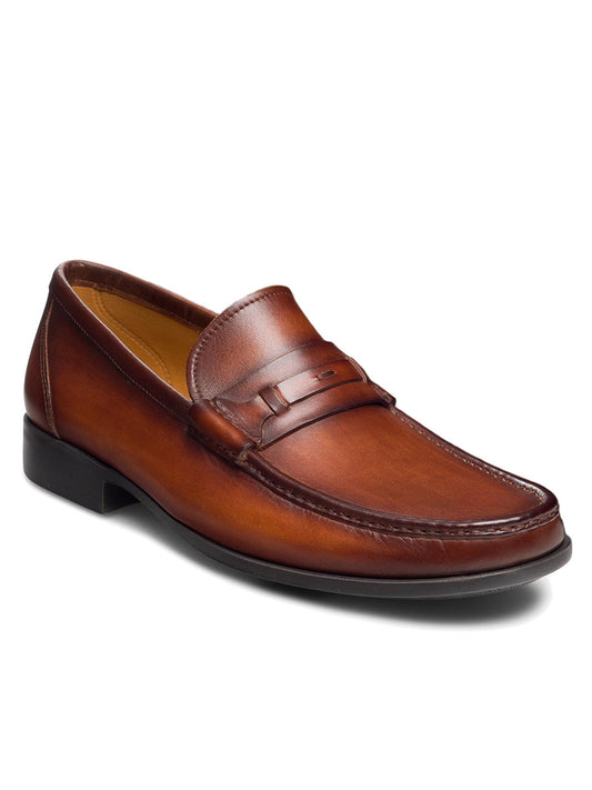 The Magnanni Daren in Cognac is a brown leather slip-on loafer with a sleek design, a low black heel, and a cushioned rubber sole, featuring a penny strap over the moccasin toe design.