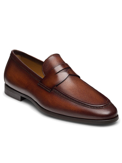 The Magnanni Dumas in Cognac penny loafer features a brown leather design with a penny strap, showcasing an elegant textured upper and a flexible rubber sole for added comfort.