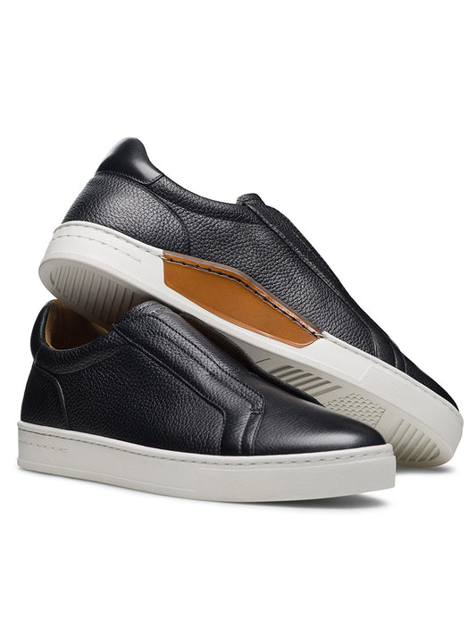 The Magnanni Gasol in Tumbled Black: black leather slip-on sneakers with hidden elastic gore, white rubber soles, and tan inner lining. One shoe stands upright while the other rests on its side to reveal the sole.