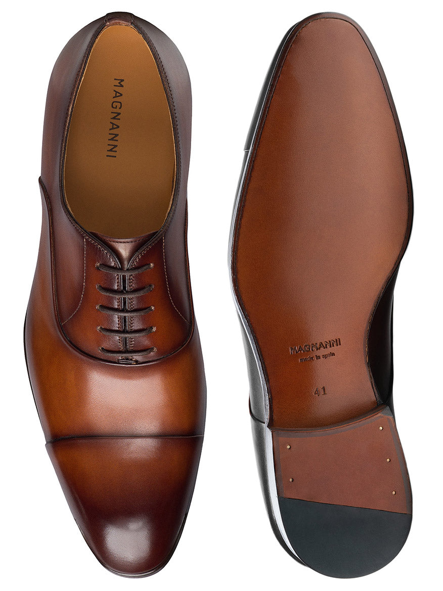 The Magnanni Naxos in Cognac is a pair of brown leather men’s formal cap toe Oxford shoes with laces, smooth polished soles, and the Magnanni logo inside—ideal for wearing with a tuxedo.