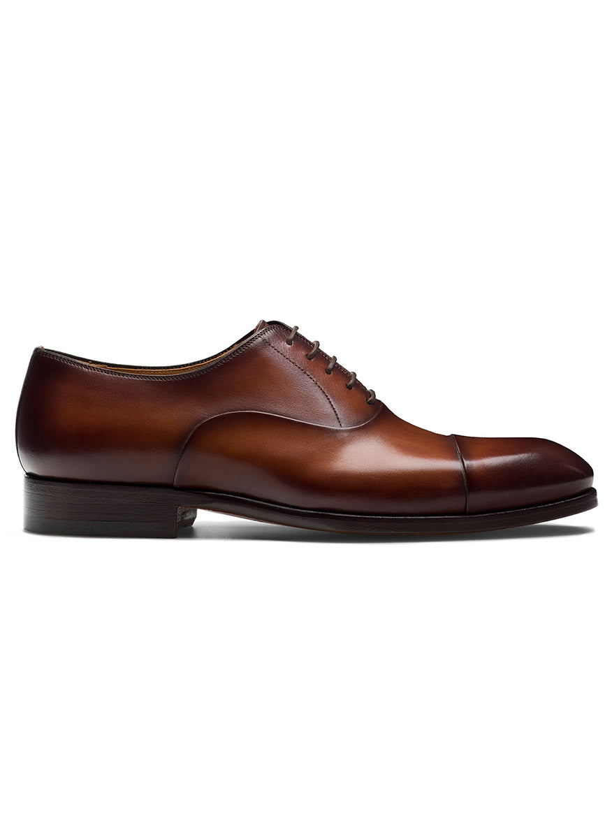 The Magnanni Naxos in Cognac is a brown leather cap toe Oxford with classic laces and a low heel, shown from the side—an elegant option for men’s formalwear.