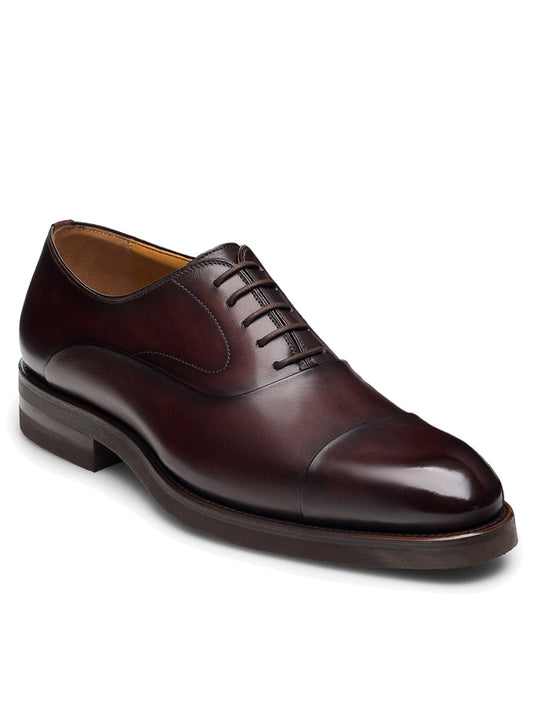 The Magnanni Omega in Midbrown is a polished brown leather Oxford shoe with a cap toe and dark sole, crafted from premium calfskin. Its neatly tied laces exude timeless elegance.