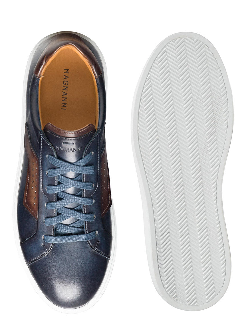 A pair of Magnanni Phoenix in Navy/Brown sneakers in blue and brown leather with white soles. The shoe on the left is viewed from above, showcasing the laces and insole, while the shoe on the right is seen from below, revealing a distinctive tread pattern and wing-inspired leather accents for a sophisticated sporty appeal.