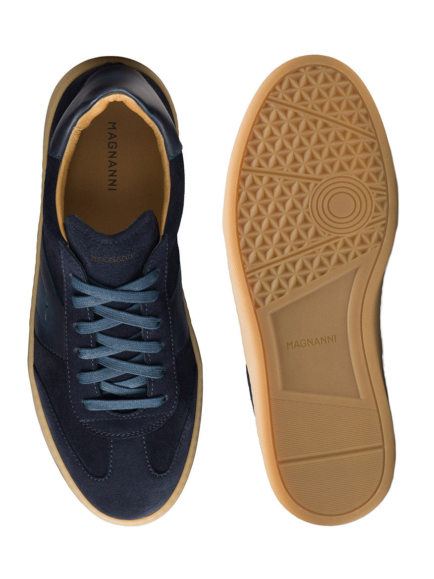 A pair of Magnanni Primera in Navy Suede, featuring dark blue premium suede uppers and beige gum soles. One shoe is displayed from the top, showcasing its stylish design, and the other from the bottom, both prominently displaying the brand name.