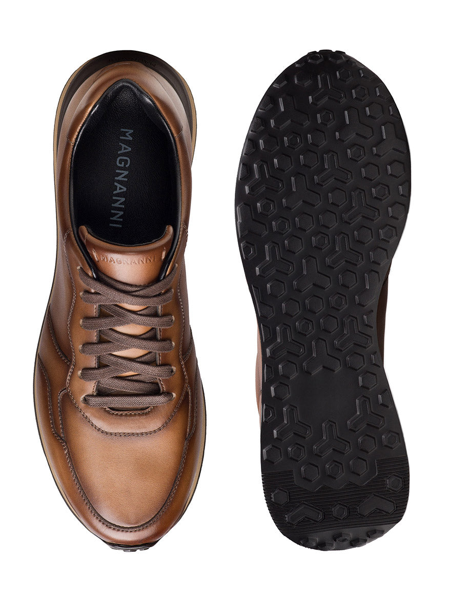 The Magnanni Syros in Brown is a brown leather running sneaker with laces, shown from above next to its black textured rubber sole. Made in Spain, the designer sneaker displays the brand name inside.