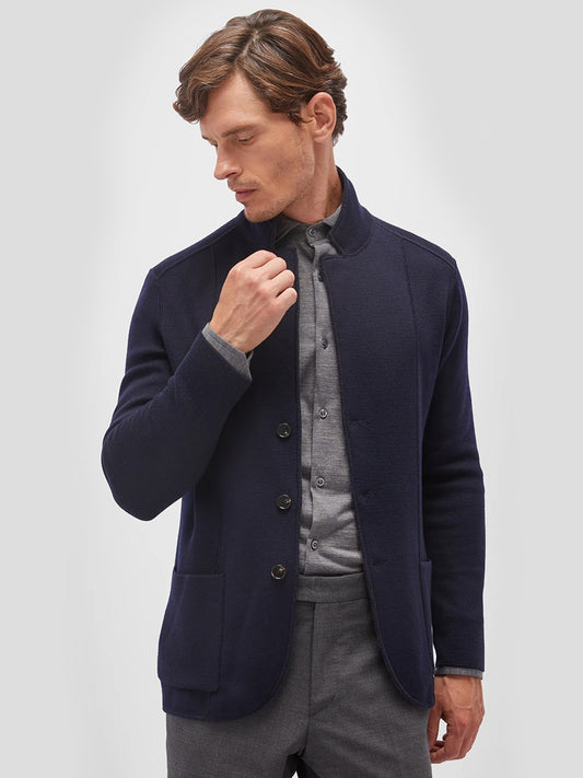 A man wearing a dark navy, Maurizio Baldassari Jamaica Reversible Wool Swacket in Navy/Charcoal over a light gray shirt and gray pants adjusts his sleeve. The background is plain white.