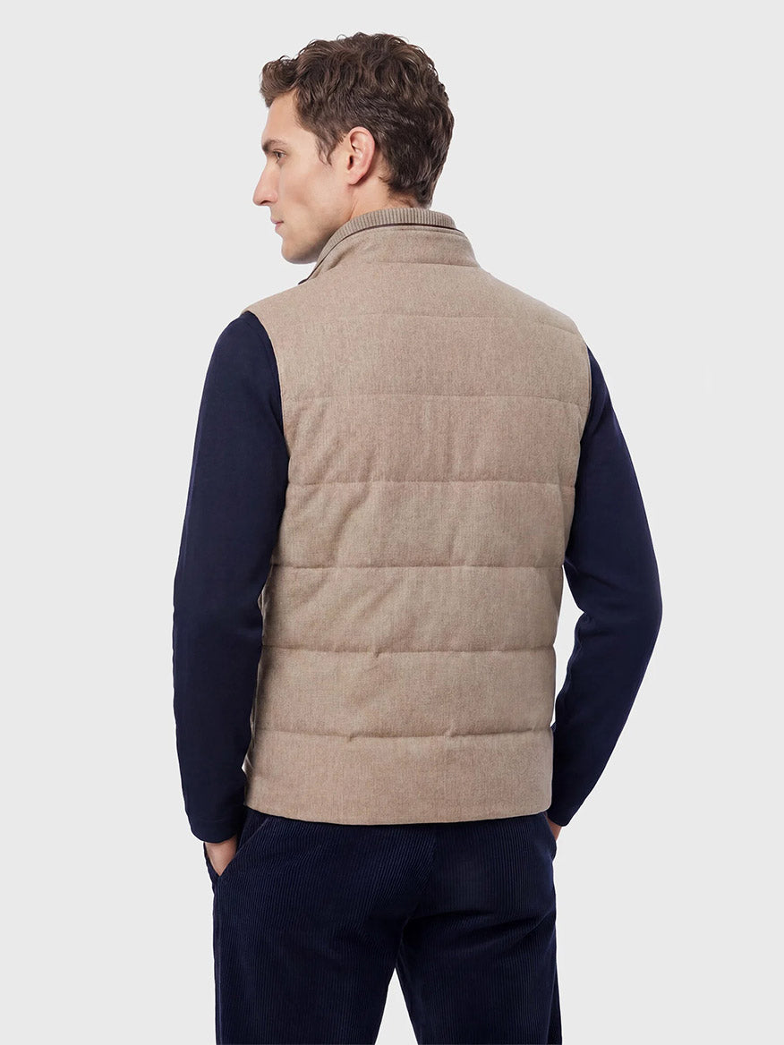 A man wearing the Maurizio Baldassari Arena Cashmere Vest in Brown Sugar—crafted in Italy—over a dark long-sleeve shirt and navy pants, stands facing away with his hands in his pockets against a plain light gray background, embodying an understated look ideal for urban living.