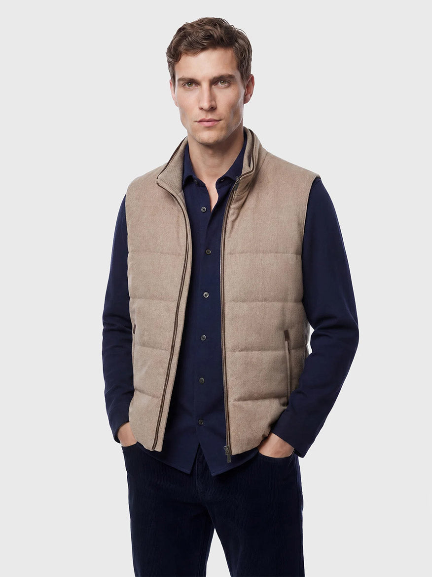 A man stands facing the camera wearing the Maurizio Baldassari Arena Cashmere Vest in Brown Sugar over a dark blue button-up shirt and dark pants against a plain light background, perfectly capturing the refined style of urban living.