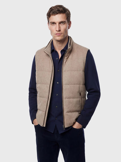 A man stands facing the camera wearing the Maurizio Baldassari Arena Cashmere Vest in Brown Sugar over a dark blue button-up shirt and dark pants against a plain light background, perfectly capturing the refined style of urban living.