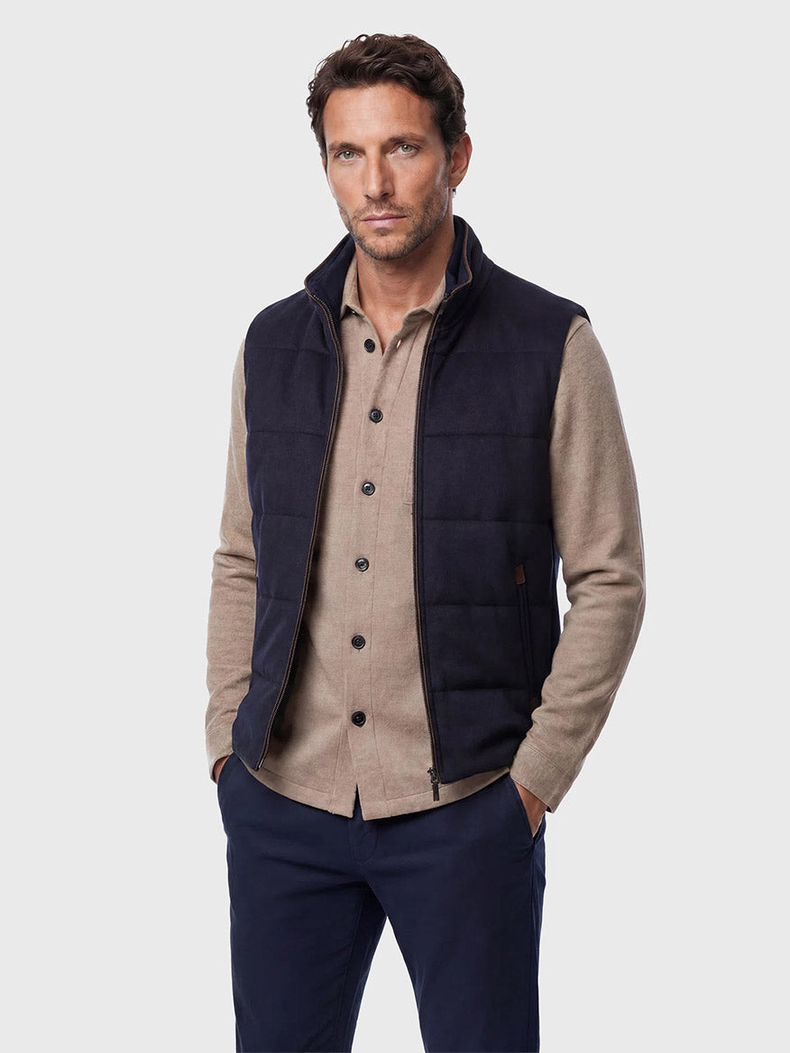 Man wearing a beige button-up shirt, Maurizio Baldassari Arena Cashmere Vest in Dark Navy, and navy pants, standing with hands in pockets against a plain light gray background—a refined look inspired by modern urban living.
