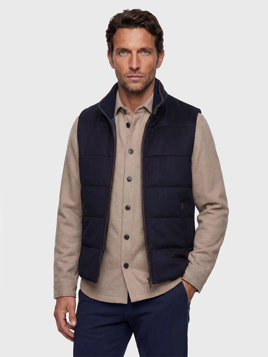 A man stands facing forward with a neutral expression against a plain light gray background, wearing a beige button-up shirt, navy pants, and the Maurizio Baldassari Arena Cashmere Vest in Dark Navy.