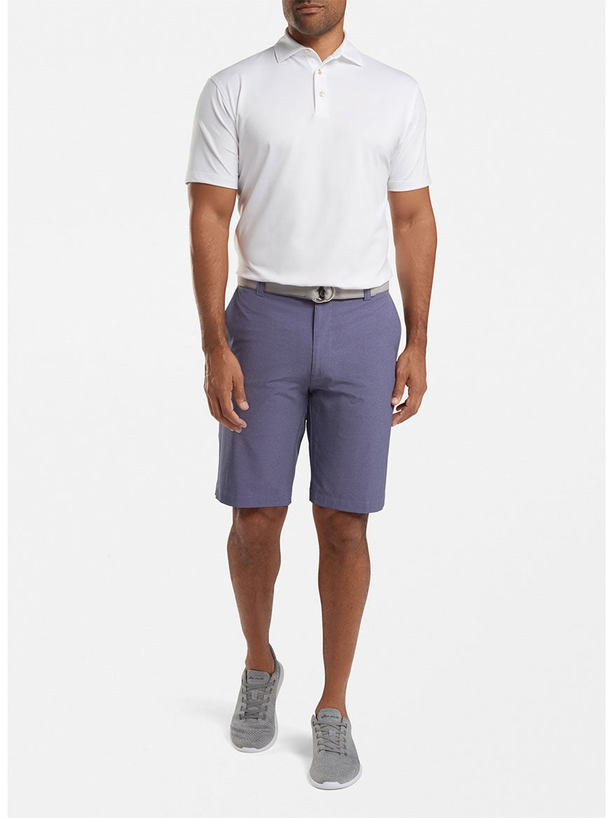 Sentence with product name: A man wearing a Peter Millar Solid Performance Jersey Polo in White [Sean Self Collar] with UPF 50+ sun protection, purple shorts, a black belt, and gray sneakers.