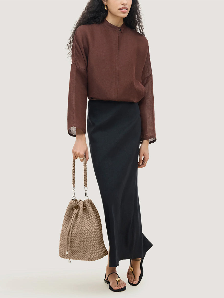 A woman wearing a brown long-sleeved shirt and black skirt carries a tan Naghedi Mercer Large Bucket in Cashmere with a braided handle.