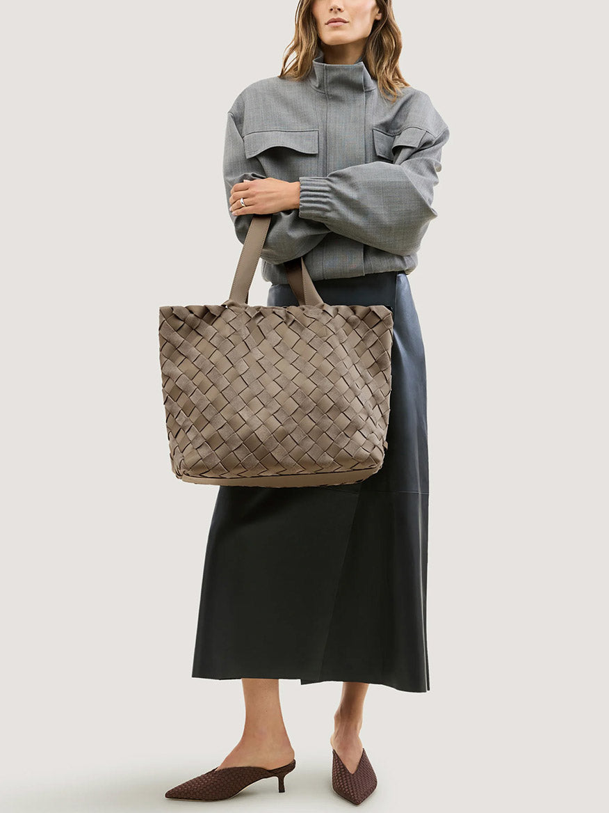 A woman holds a Naghedi Como Medium Tote Suede & Neoprene in Bluff, a medium-sized, handwoven Italian suede tote bag in a muted taupe color with two shoulder straps.