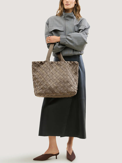 A woman holds a Naghedi Como Medium Tote Suede & Neoprene in Bluff, a medium-sized, handwoven Italian suede tote bag in a muted taupe color with two shoulder straps.