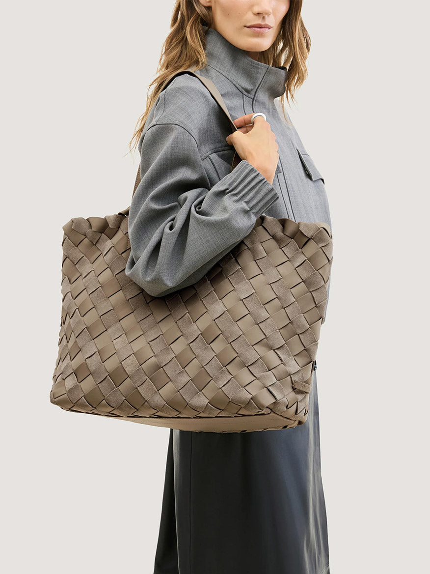 A person carrying a large, taupe handwoven Naghedi Como Medium Tote Suede & Neoprene in Bluff over their shoulder.