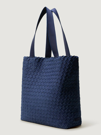The Naghedi Como Shopper Tote in Denim, a navy blue handwoven bag with two long handles, stands upright against a plain light background.