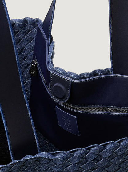 Close-up of the Naghedi Como Shopper Tote in Denim. This unisex, blue handwoven bag features a textured exterior, fabric straps, snap button closure, and a zippered inner pocket.