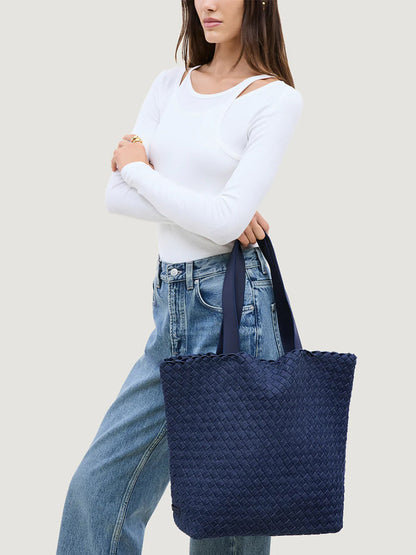 A woman stands against a plain light background, wearing a white long-sleeve top and blue jeans while carrying the Naghedi Como Shopper Tote in Denim over her arm.