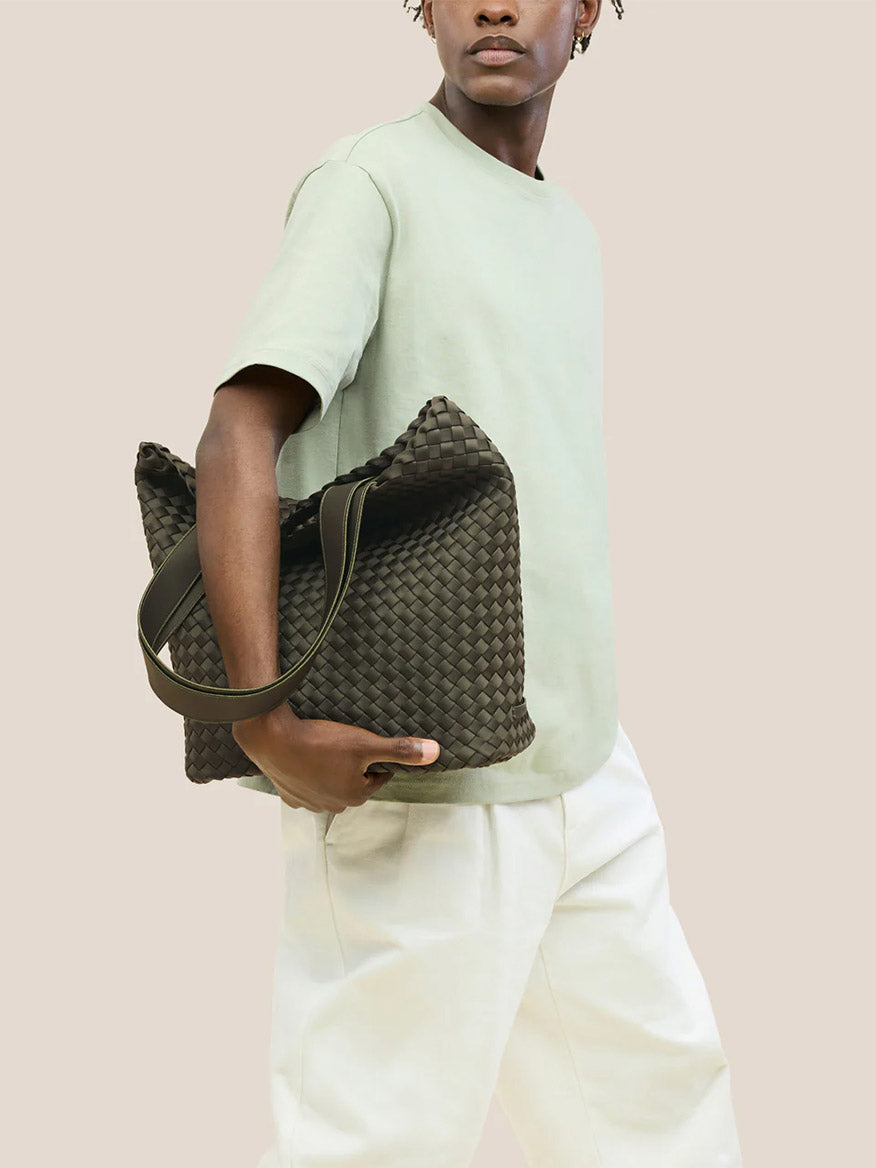 A person in a light green t-shirt and white pants holds the Naghedi Como Shopper Tote in Olive against a plain beige background.
