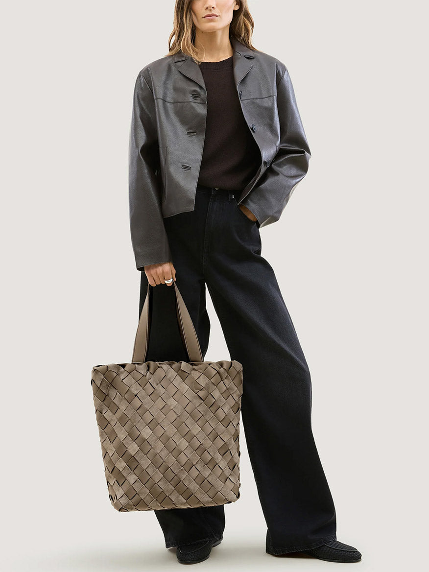 A person wearing a dark jacket and jeans holds a taupe woven Naghedi Como Shopper Tote Suede & Neoprene in Bluff.