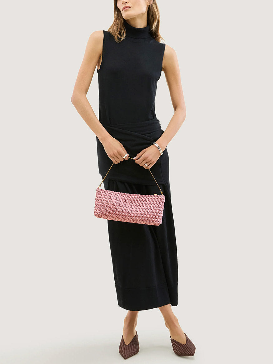 A person wearing a black sleeveless dress holds a Naghedi Gala Clutch Satin in Flor, a woven ballet pink clutch with a thin gold chain strap.