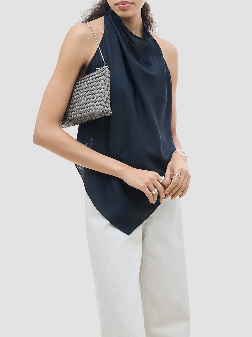 A person wearing a dark blue halter top and white pants holds a gray woven Naghedi Gala Clutch Satin in Graphite under their arm.