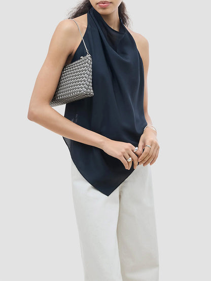 A person wearing a dark blue halter top and white pants holds a gray woven Naghedi Gala Clutch Satin in Graphite under their arm.