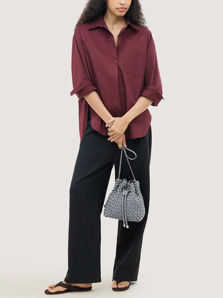 A person wearing a maroon button-down shirt and black trousers holds a gray Naghedi Gramercy Bucket Double Weave in Marina handwoven bucket bag with a drawstring closure.