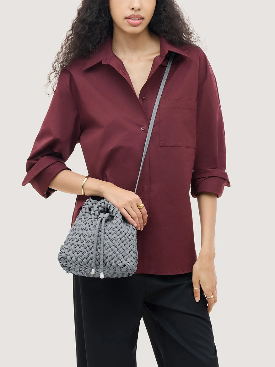 A person wearing a maroon button-down shirt and carrying a grey handwoven Naghedi Gramercy Bucket Double Weave in Marina with a drawstring closure and shoulder strap.