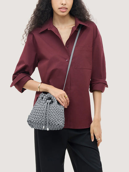 A person wearing a maroon button-down shirt and carrying a grey handwoven Naghedi Gramercy Bucket Double Weave in Marina with a drawstring closure and shoulder strap.