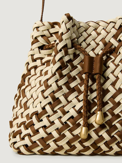 Close-up of the Naghedi Gramercy Bucket Double Weave in Carmel, showcasing its beige and brown interlaced pattern and two braided drawstrings finished with gold-tone metal tips.