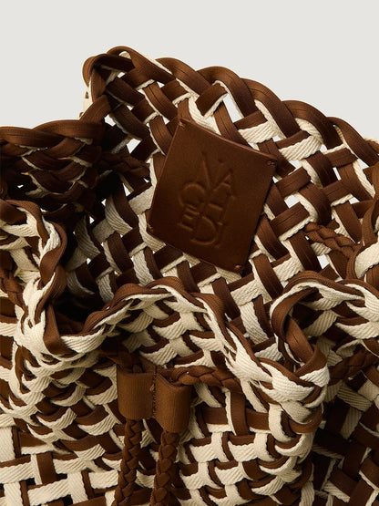 Close-up of the Naghedi Gramercy Bucket Double Weave in Carmel, featuring a woven brown and white design and a brown leather patch with embossed text.