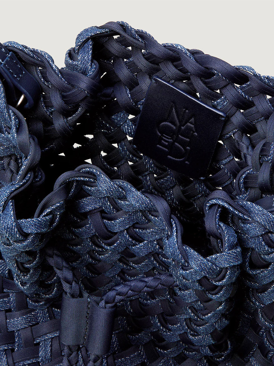 Close-up of the Naghedi Gramercy Bucket Double Weave in Indigo, a navy handwoven handbag with a textured pattern and a black tag embossed with "NAGHEDI" branding.