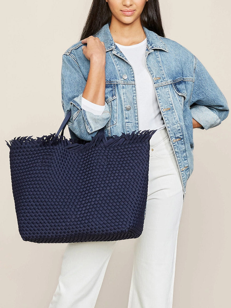 A woman modeling a denim jacket and white pants while carrying a Naghedi Havana Beach Tote in Solid Ink Blue Fringe.