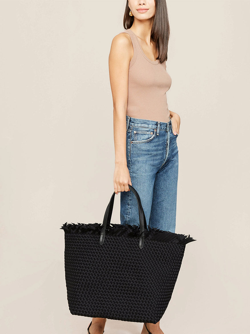 A woman in a beige tank top and blue jeans holding a Naghedi Havana Large Travel Tote in Solid Onyx Fringe with vegan leather handles and fringe detail.