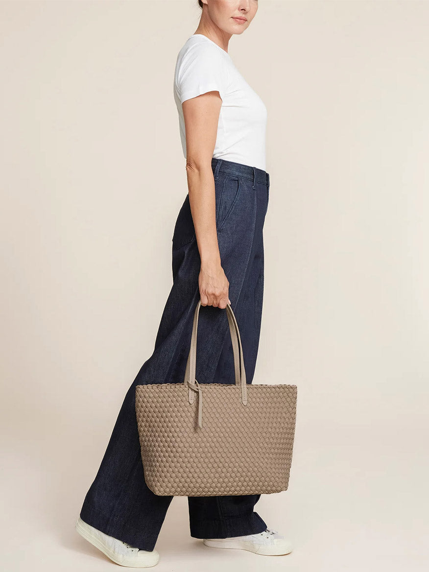 A person wearing a white t-shirt and wide-leg jeans is holding the Naghedi Jetsetter Small Tote in Solid Cashmere, which is crafted from handwoven neoprene.