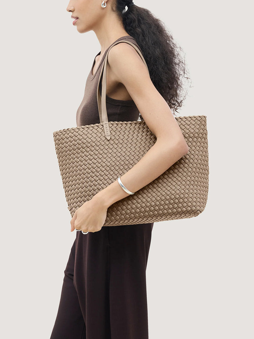 A person holds a large, beige handwoven neoprene Naghedi Jetsetter Small Tote in Cashmere under their arm.