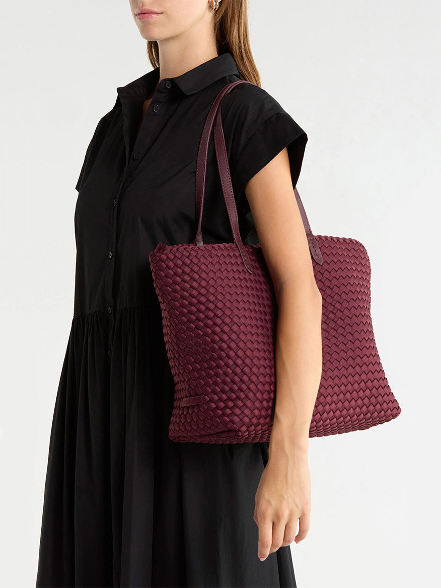A person wearing a black dress carries a deep red Naghedi Jetsetter Small Tote over their shoulder.