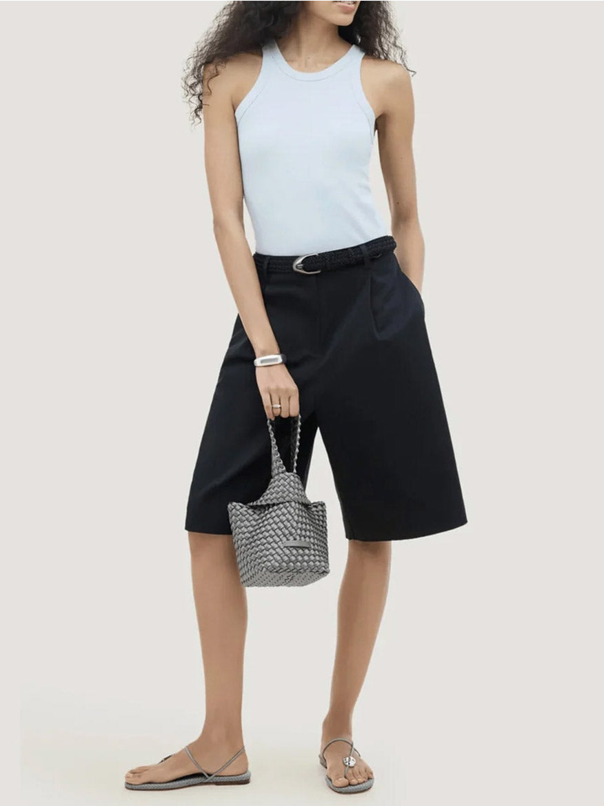 A person wearing a light blue tank top and dark shorts holds a small, grey Naghedi Kyoto Clutch Satin in Graphite.