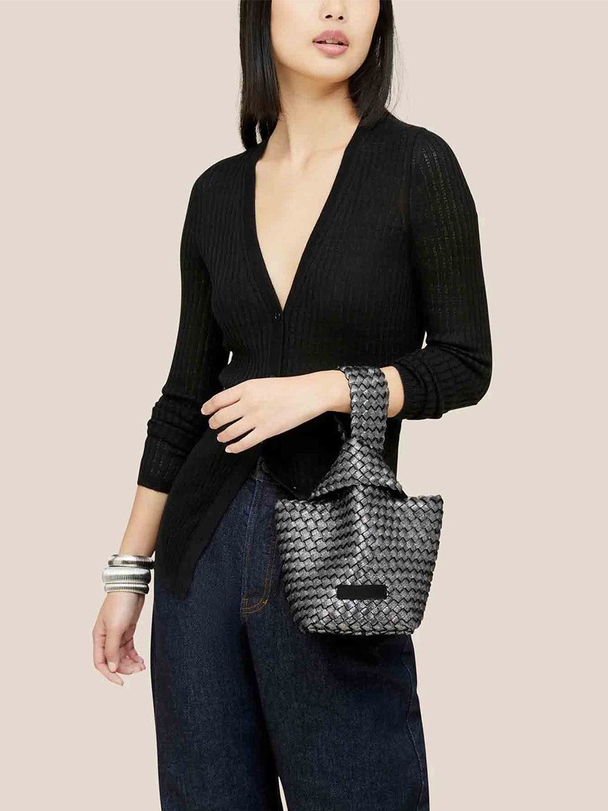 A woman in a black sweater and jeans holds the Naghedi Kyoto Clutch in Metallic Titanium, perfect for evening outfits.