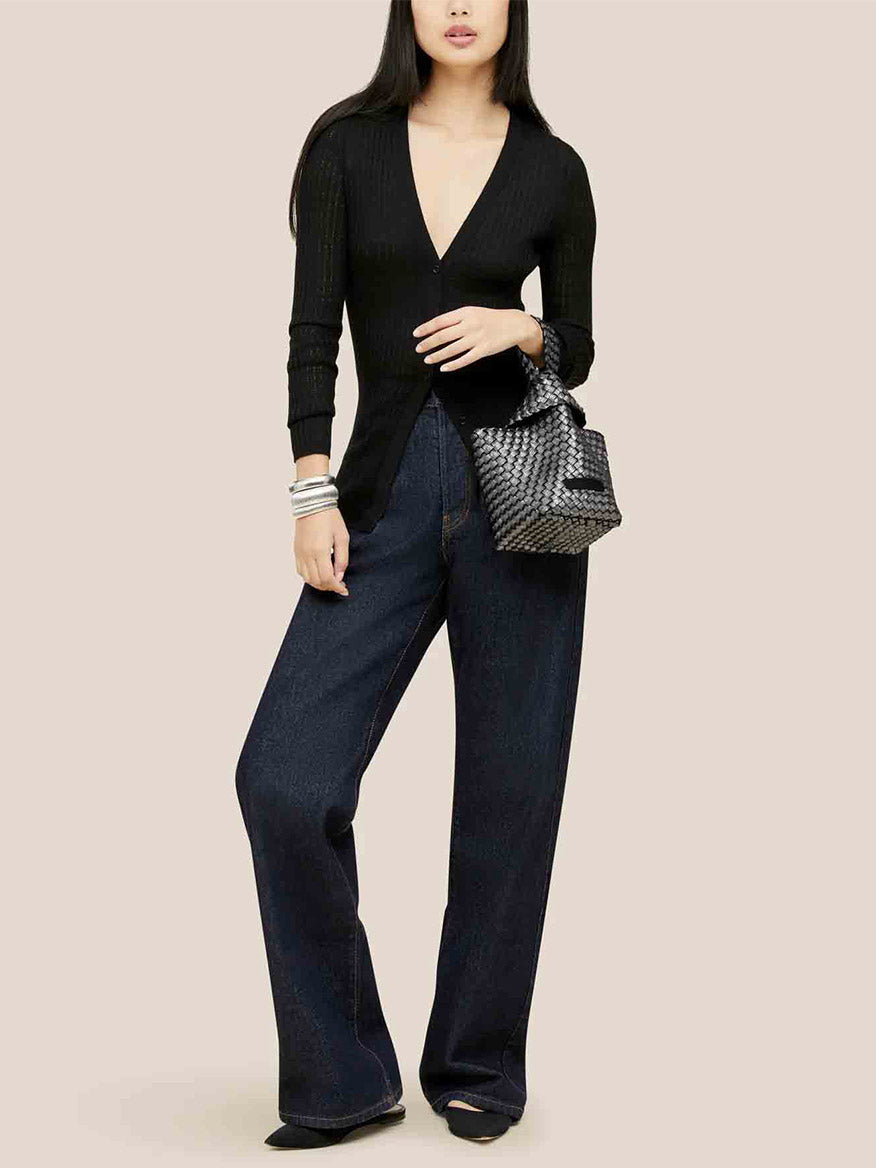 A woman wearing a black V-neck top and wide-leg jeans, holding a Naghedi Kyoto Clutch in Metallic Titanium.