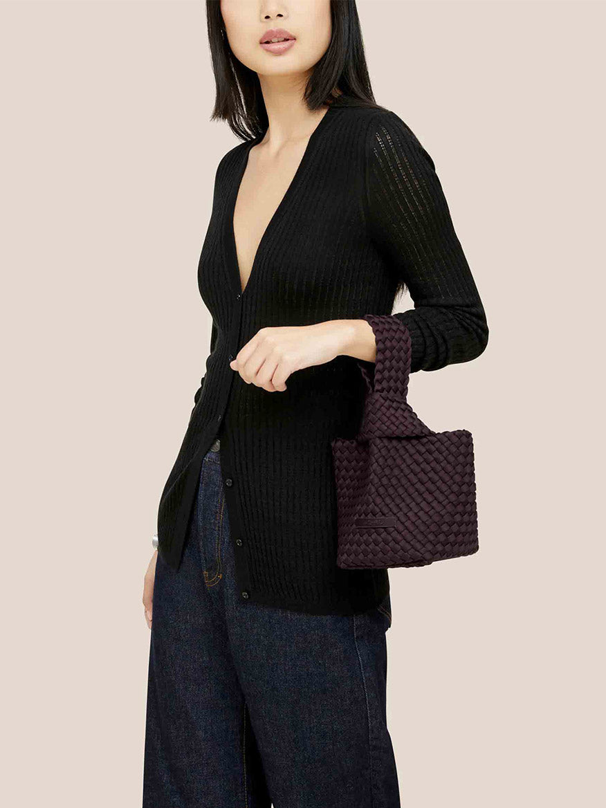 A person wearing a black cardigan and jeans holds a Naghedi Kyoto Clutch in Solid Aubergine, perfect for complementing evening outfits.